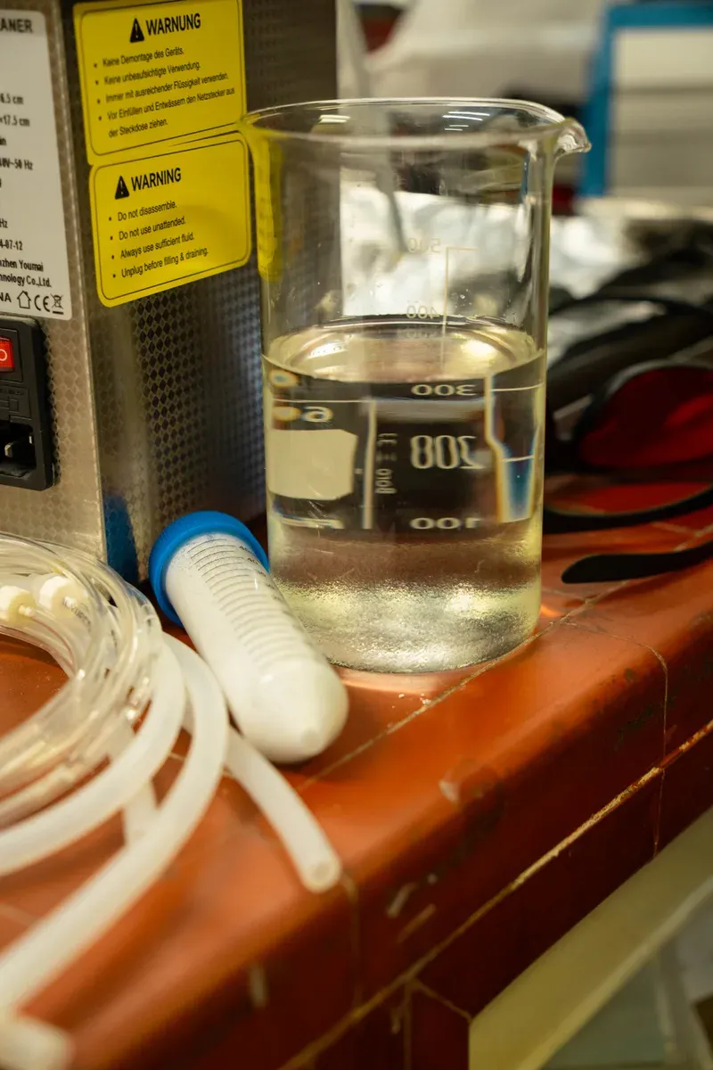 A glass beaker of pale liquid on a lab bench beside a graduated tube and coiled tubing, next to an appliance carrying German and English safety warning labels.