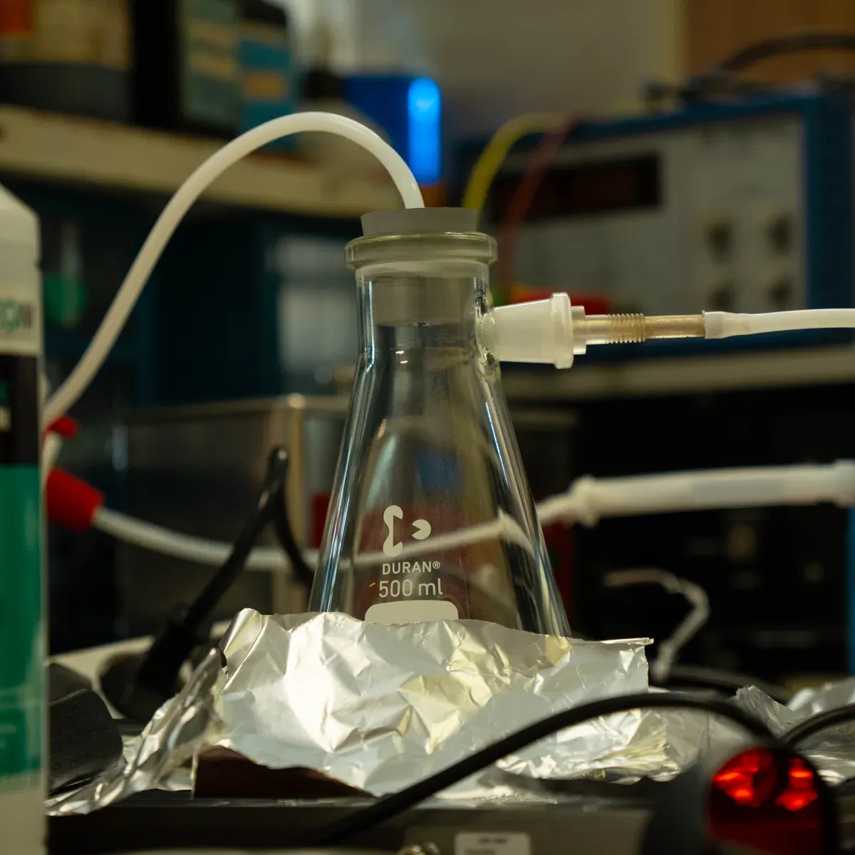 A 500 mL Duran Erlenmeyer flask stoppered and wired into lab equipment, a white gas line running from its top and an electrode fitting threaded into its side, sitting on a bed of aluminum foil.