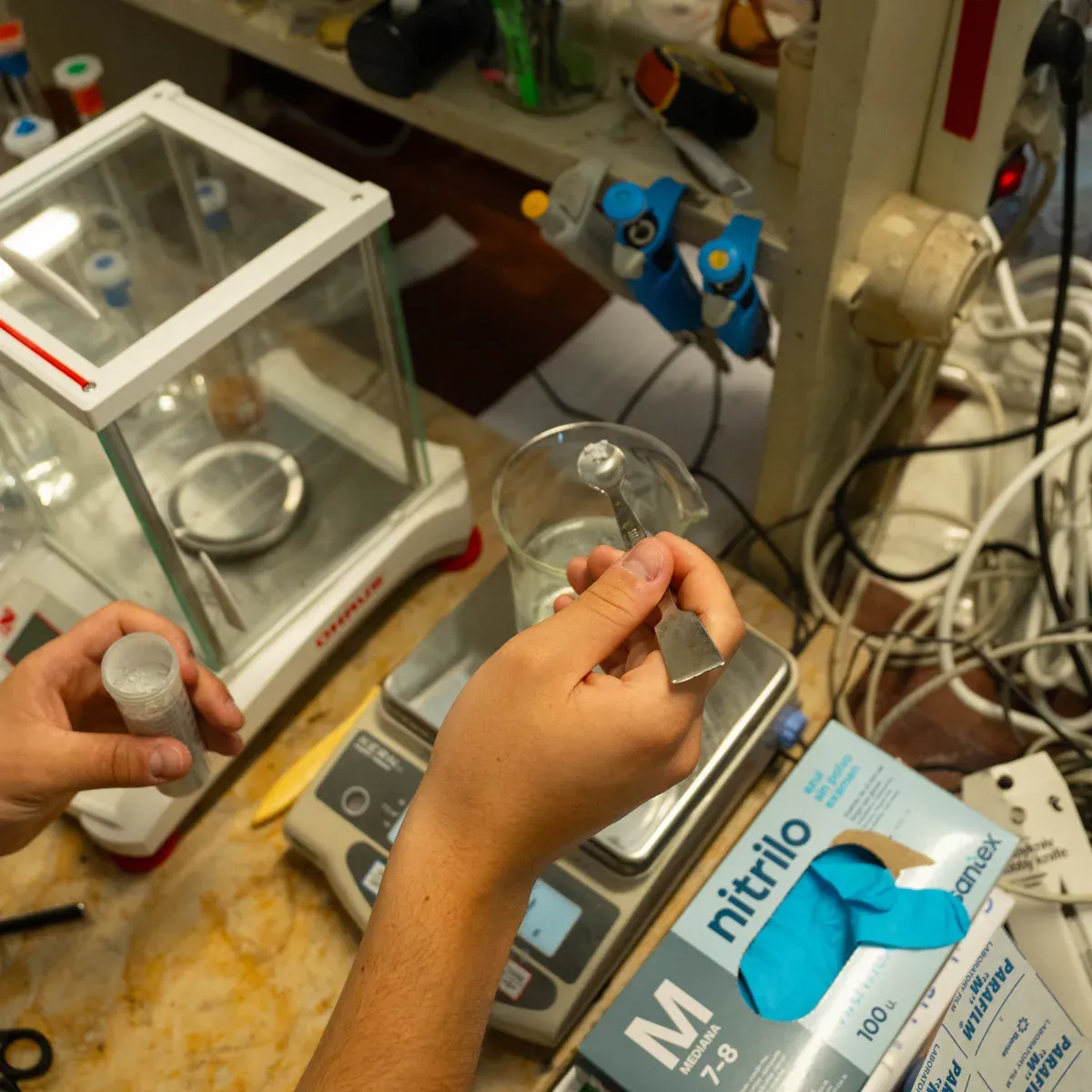 Overhead view of two hands weighing a white powder on a small metal spatula over a beaker, beside an analytical balance in its glass draft shield.