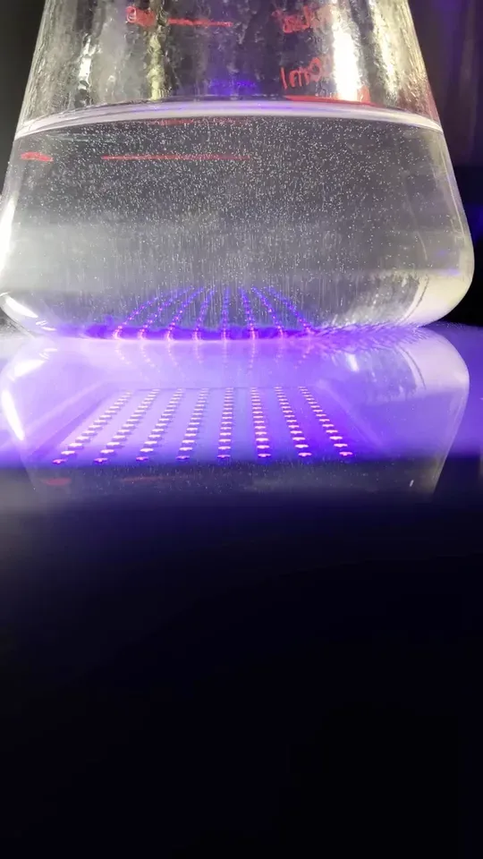 A graduated flask of water bubbling on a violet UV LED panel in a darkened room, gas rising to the surface.