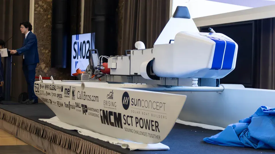 A presenter at a podium beside Técnico Solar Boat's new hydrogen-powered catamaran on stage, a screen behind reading 'SM02'; the hull is lettered with event sponsor names including CalliBros.com, SunConcept Solar Boat Builders, and INCM SCT Power, 'Solving Challenges Together'.